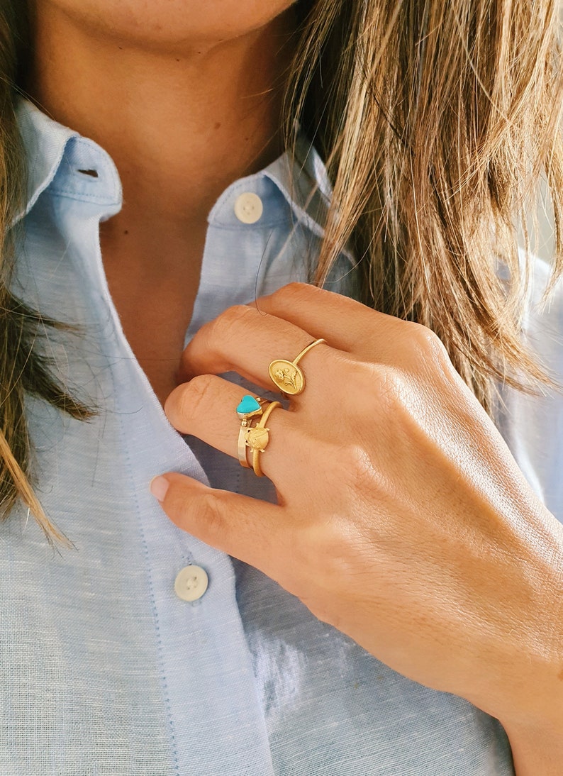 Pu&ograve; includere: Una mano con tre anelli d'oro. Gli anelli presentano un cuore turchese, un piccolo cuore d'oro e un ovale pi&ugrave; grande con un design in rilievo.