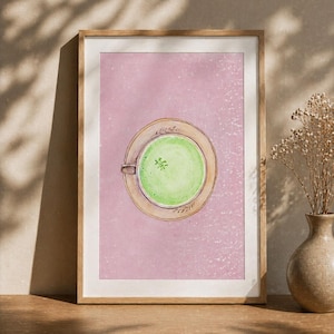 May include: Watercolor print of a matcha latte in a light brown cup and saucer, set against a pink background. The artwork is framed in a light brown wooden frame. A vase with dried flowers is on the right.
