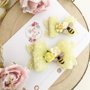 May include: Two yellow glitter hair bows with a bee and flower embellishments. The bows are on a white card with the text "Roses & Bows" and a website address.
