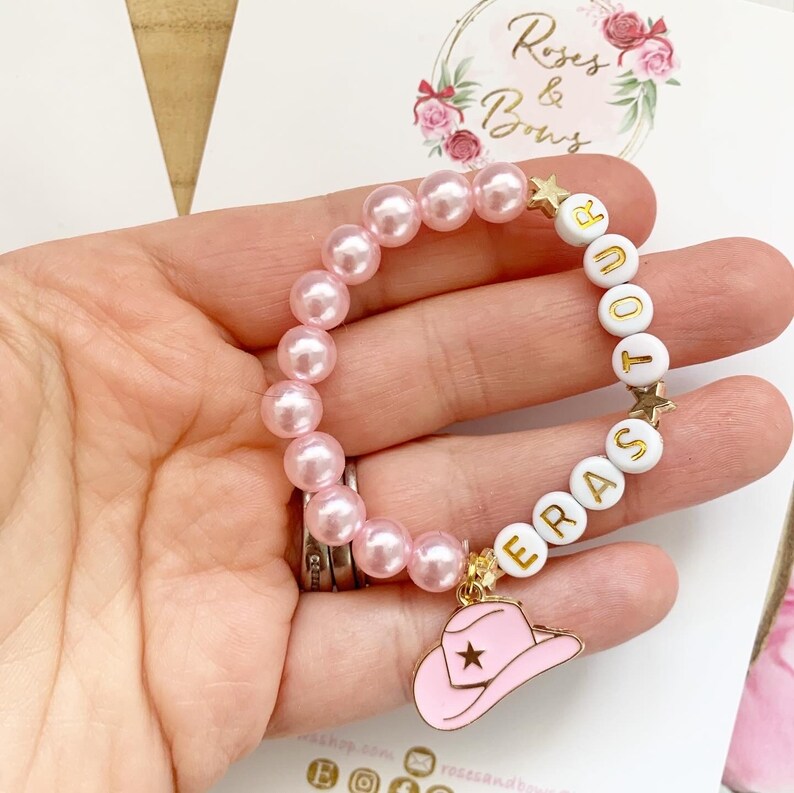 May include: Pink pearl bracelet with white letter beads spelling "ERAS TOUR" and a gold charm of a pink cowboy hat. The bracelet is held in a hand, with a logo in the background that says "Roses & Bows".