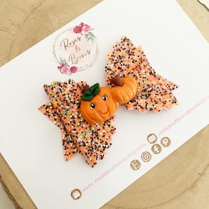 Könnte beinhalten: Handgefertigte orangefarbene Glitzer-Haarschleife mit einem lächelnden Kürbisgesicht. Die Schleife ist mit schwarzen, weißen und goldenen Glitzer verziert. Die Schleife liegt auf einer weißen Karte mit dem Text "Roses & Bows".