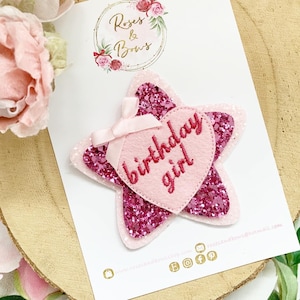 Puede incluir: Estrella de purpurina rosa con un corazón de fieltro rosa que dice "birthday girl" en bordado rojo.