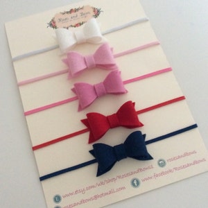 May include: Set of five felt hair bows in white, pink, light pink, red, and navy blue. Each bow is attached to a nude elastic headband.