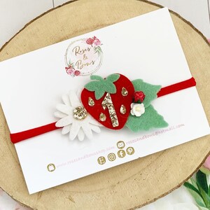 May include: A red elastic headband featuring a felt strawberry with glitter accents, a white daisy, and green leaves. The strawberry has a glitter number one. The headband is on a white card with the text "Roses & Bows".