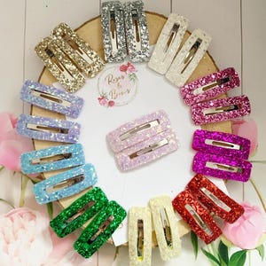 Könnte beinhalten: Ein Set mit 10 Paaren glitzernden Haarklammern in verschiedenen Farben, darunter Silber, Gold, Weiß, Rosa, Lila, Rot, Grün und Blau. Die Klammern sind in einem Kreis auf einer Holzoberfläche angeordnet. Der Text "Roses & Bows" ist in der Mitte des Kreises sichtbar.