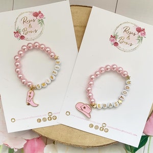 May include: Two pink beaded bracelets with white letter beads spelling "Swiftie" and "Eras Tour". Each bracelet has a charm: a pink cowboy boot and a pink cowboy hat. The bracelets are on white cards.