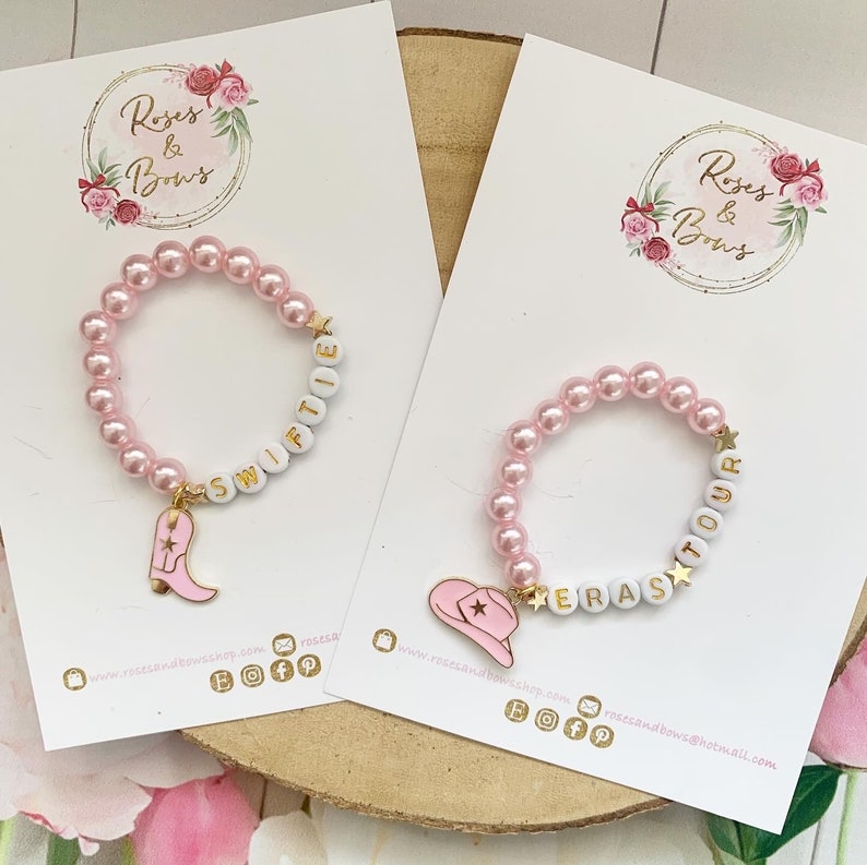 May include: Two pink beaded bracelets with white letter beads spelling "Swiftie" and "Eras Tour". Each bracelet has a charm: a pink cowboy boot and a pink cowboy hat. The bracelets are on white cards.