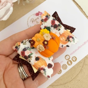 Könnte beinhalten: Handgefertigte Haarspange zum Thema Herbst mit einer orangefarbenen Kürbis-Mitte und floralen Verzierungen. Die Schleife hat Stofflagen mit einem Blumen- und Kürbismuster sowie einen dunkelroten und goldenen Akzent. Für einen festlichen Herbst-Look.