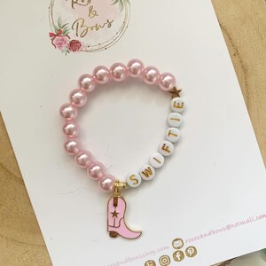 May include: Pink pearl bead bracelet with white letter beads spelling "Swiftie" and a gold star charm. A pink cowboy boot charm hangs from the bracelet. The bracelet is on a white surface with the "Roses & Bows" logo.