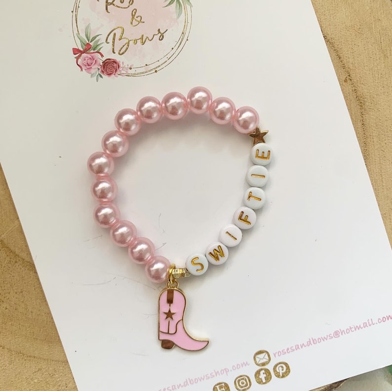 May include: Pink pearl bead bracelet with white letter beads spelling "Swiftie" and a gold star charm. A pink cowboy boot charm hangs from the bracelet. The bracelet is on a white surface with the "Roses & Bows" logo.