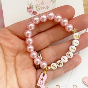 May include: Pink pearl bead bracelet with white letter beads spelling "SWIFT" and a gold star charm. A pink and gold cowboy boot charm hangs from the bracelet. The bracelet is a charming accessory.