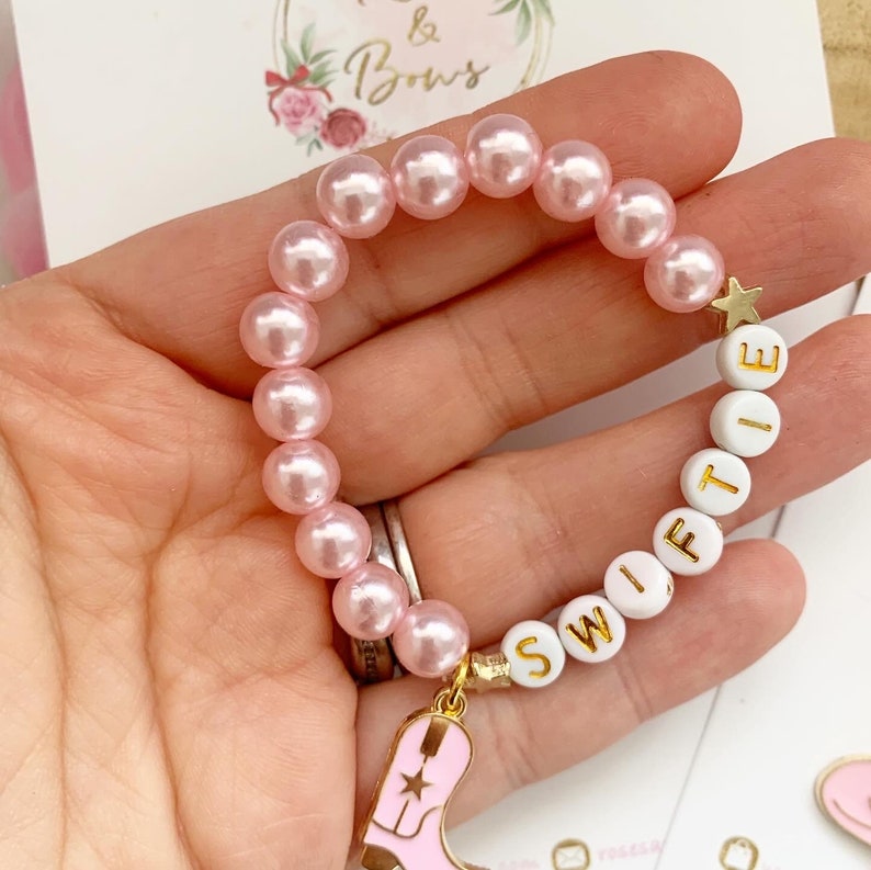 May include: Pink pearl bead bracelet with white letter beads spelling "SWIFT" and a gold star charm. A pink and gold cowboy boot charm hangs from the bracelet. The bracelet is a charming accessory.
