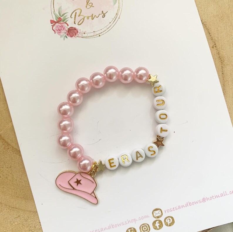 May include: Pink pearl bracelet with white letter beads spelling "ERAS TOUR". The bracelet features a pink cowboy hat charm and gold star accents. The bracelet is on a white card with the "Roses & Bows" logo.