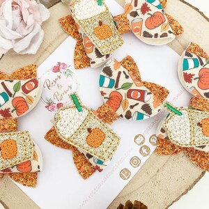May include: A collection of autumn-themed hair bows with glitter detailing. Some bows feature a pumpkin spice latte design with an embroidered pumpkin. Other bows have a pattern of pumpkins, leaves, and coffee cups. The bows are on a white surface with the text "Roses & Bows".