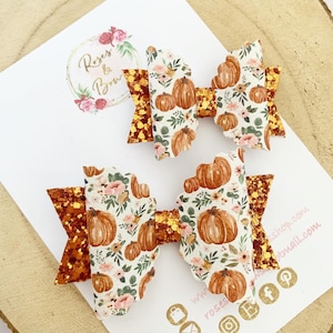 Peut inclure: Deux nœuds décoratifs pour les cheveux. Les nœuds présentent un motif de citrouille et floral sur fond blanc, avec des accents orange pailletés. Les nœuds sont sur une carte blanche avec le texte "Roses & Bows".
