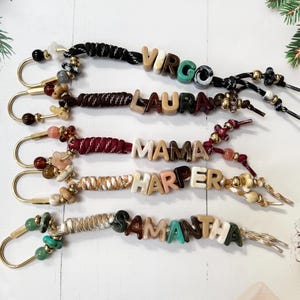 May include: Five keychains featuring gold-toned U-shaped clasps, braided nylon cords in various colours, and assorted beads. The letter beads spell out VIRGO, LAURA, MAMA, HARPER, and SAMANTHA in a mix of neutral, brown, green, and teal tones.