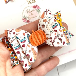 May include: A handmade hair bow with a pumpkin centrepiece. The bow features a white background with autumn-themed patterns, including pumpkins, leaves, and mugs. The bow has glitter accents in orange, gold, and brown.
