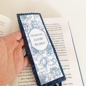May include: A rectangular bookmark with a dark blue glittery border and a white centre panel featuring blue line-art floral illustrations. The text "Dearest Gentle Reader" is printed in the centre within a decorative frame, with a small blue ribbon loop.