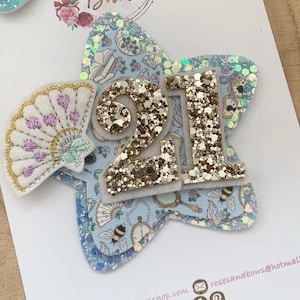 Puede incluir: Accesorio para el cabello en forma de estrella con un fondo azul claro con abejas, relojes y otros diseños. El número "21" se muestra prominentemente en purpurina dorada. Un pequeño abanico está adjunto al costado.