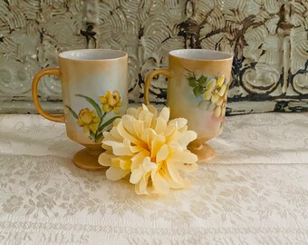May include: Two vintage ceramic mugs with floral designs. The mugs are beige with yellow flowers and green leaves. The mugs have a pedestal base and are sitting on a white lace tablecloth.