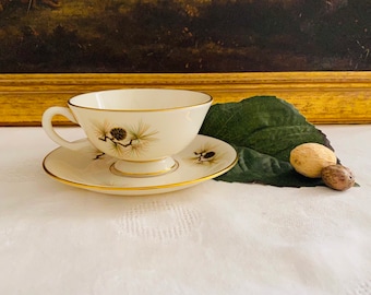 May include: A white teacup and saucer with a gold rim and a pine cone design. The cup and saucer are on a white tablecloth with a green leaf and two brown nuts.