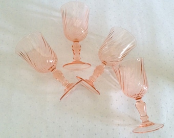 May include: Four pink glass wine glasses with a swirl design. The glasses are arranged in a circle on a white surface.