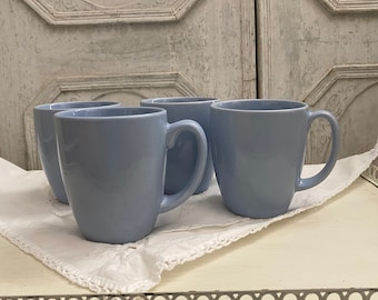 May include: Four light blue ceramic mugs with handles. The mugs are arranged in a semi-circle on a white doily.