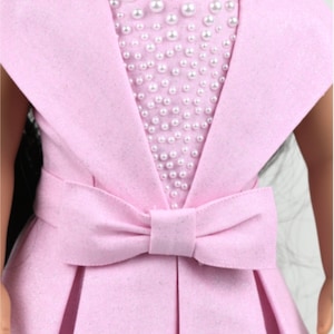 May include: A pink dress with a pearl embellishment on the bodice and a large bow at the waist. The dress is designed for a doll.