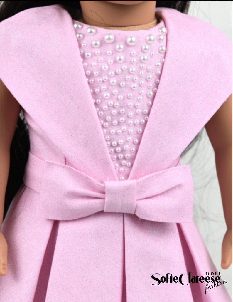 May include: A pink dress with a pearl embellishment on the bodice and a large bow at the waist. The dress is designed for a doll.