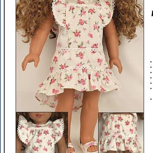 Op de afbeelding: Een witte jurk met roze bloemenprint en kantversiering. De jurk heeft een hoge halslijn en korte mouwen met ruches. De jurk is gestyled op een pop met krullend bruin haar.