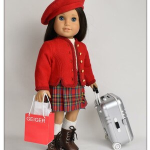 May include: A red beret, red jacket, and plaid skirt outfit for a doll. The doll is holding a red shopping bag with the word "GEIGER" printed on it and a silver suitcase with wheels.