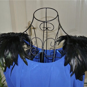 Puede incluir: Un top azul sin mangas con hombreras de plumas negras. El top está en un maniquí de metal negro.