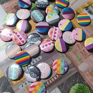 Puede incluir: Una colección de botones redondos coloridos con varios diseños, incluyendo corazones, rayas, purpurina y cinta washi. Los botones están dispuestos sobre una superficie de madera con texto en francés.
