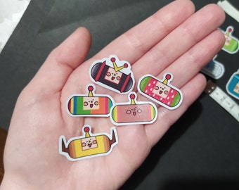 以下が含まれることがあります： 小さなアンテナが付いた、笑顔の長方形のキャラクターが描かれた、カラフルで漫画風のステッカーのコレクション。ステッカーには、ストライプ、王冠、フルーツパターンなど、さまざまなデザインがあります。ステッカーは手に持たれています。