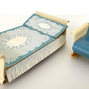 May include: A miniature metal dollhouse bed and chair set. The bed features a cream-colored frame with a blue and white floral patterned cover. The matching chair has a solid blue seat and backrest with cream-colored armrests and base supports.