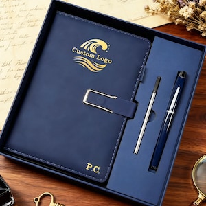 Może przedstawiać: Granatowy zestaw notatnika i długopisu w pudełku prezentowym. Notatnik ma złote zapięcie oraz napis "Custom Logo" i "P.C.". Zestaw zawiera długopis i pióro wieczne. Widoczny jest również zabytkowy klucz, butelka z atramentem i lupa.