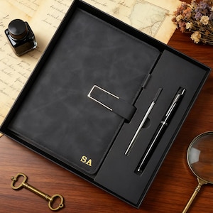 Puede incluir: Un diario de cuero negro con cierre plateado y las iniciales "SA" en dorado, presentado en una caja negra con dos bolígrafos. Se muestra sobre una superficie de madera con una llave y una lupa vintage.