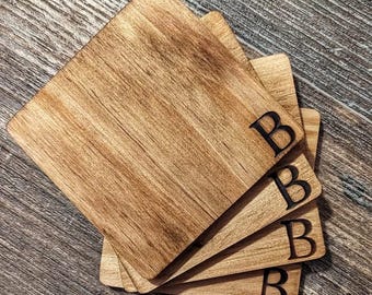 Puede incluir: Un conjunto de cuatro posavasos cuadrados de madera con vetas naturales. Cada posavasos tiene la letra "B" grabada en negro. Los posavasos están colocados sobre una superficie de madera.
