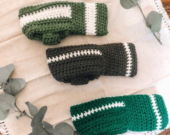 May include: Three crocheted green and white striped fingerless gloves. The gloves are made with a white yarn and a green yarn. The gloves are laid out on a white fabric with a fringe border.