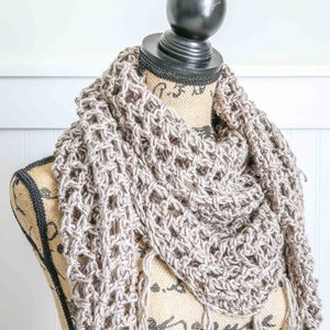 May include: A beige crochet scarf with fringe on a  mannequin. The scarf is made with a simple stitch pattern and has a delicate, airy feel.
