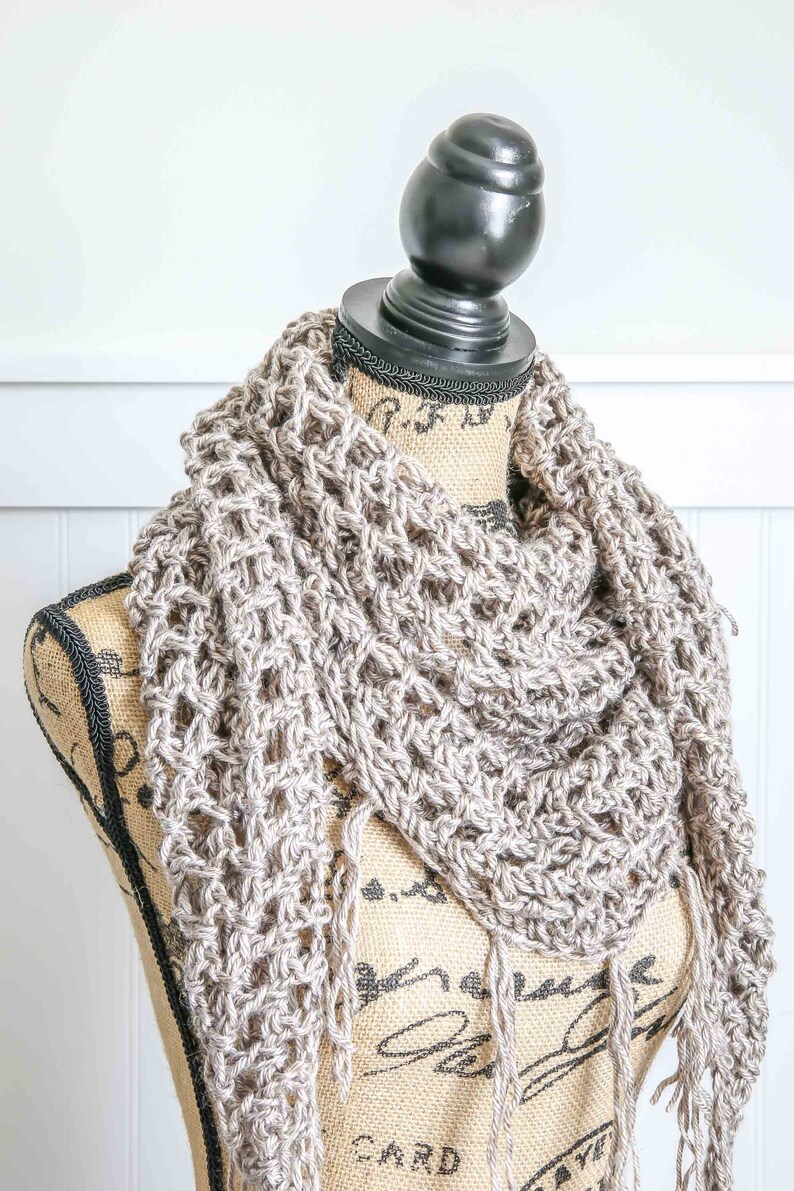 May include: A beige crochet scarf with fringe on a  mannequin. The scarf is made with a simple stitch pattern and has a delicate, airy feel.