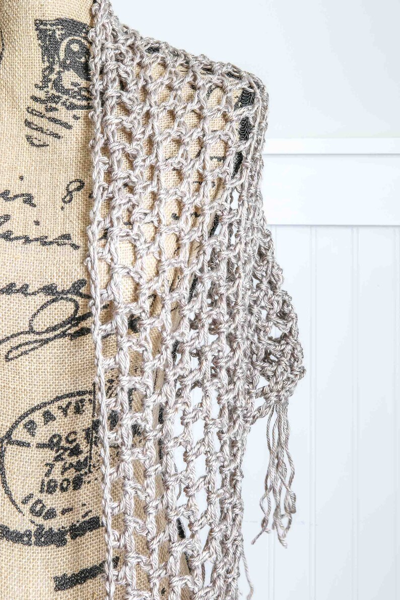 May include: A silver crochet shawl with a delicate openwork pattern and fringe trim. The shawl is draped over a mannequin.