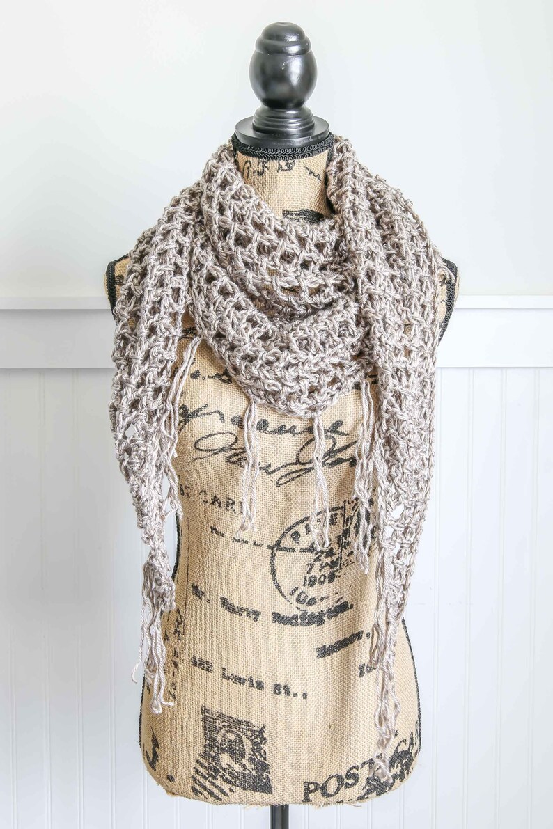 May include: A crocheted scarf in a light brown color with fringe on the ends. The scarf is draped over a burlap mannequin.