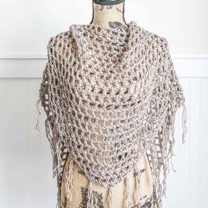 May include: A crocheted shawl with a delicate openwork pattern and fringe trim. The shawl is displayed on a mannequin and is a light beige color.