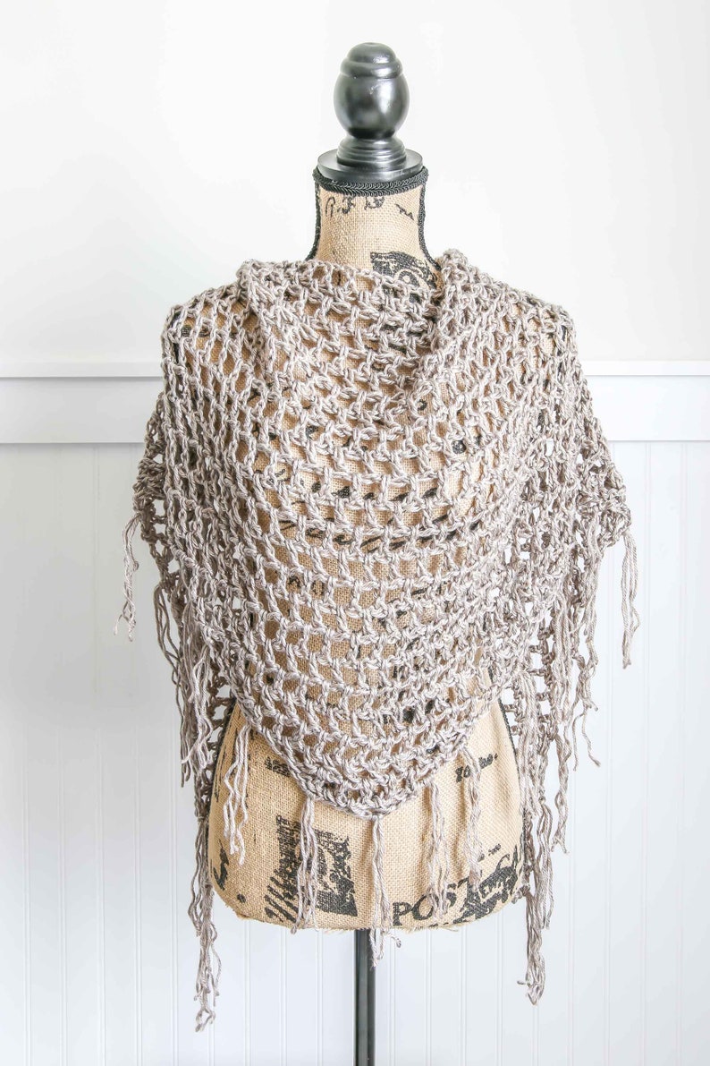 May include: A crocheted shawl with a delicate openwork pattern and fringe trim. The shawl is displayed on a mannequin and is a light beige color.