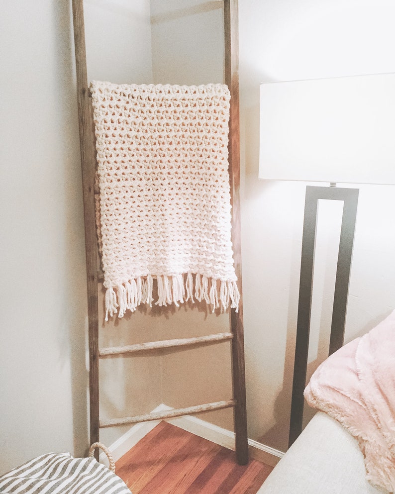 May include: A white knitted throw blanket with fringe hanging on a wooden ladder leaning against a wall.