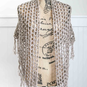 May include: A crocheted shawl in a light brown color with fringe on the edges. The shawl is draped over a mannequin.