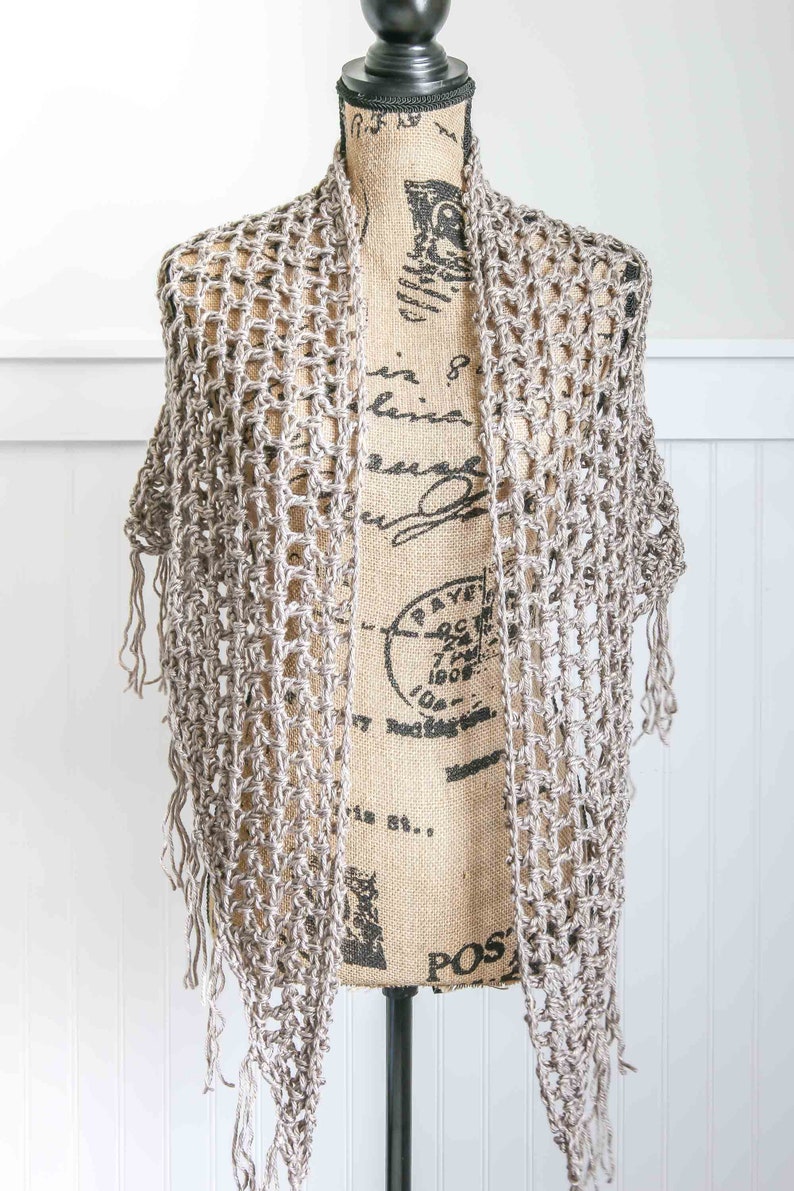 May include: A crocheted shawl in a light brown color with fringe on the edges. The shawl is draped over a mannequin.