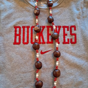May include: A grey t-shirt featuring the red text BUCKEYES and a red Nike swoosh logo, layered with a long necklace made of large brown buckeye seeds interspersed with small red and white beads arranged in a repeating pattern along the string.