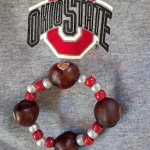 May include: A circular bracelet featuring four smooth, dark brown buckeye nuts interspersed with alternating red and silver spherical beads. The bracelet is positioned against a grey fabric background displaying the Ohio State logo in red and black.